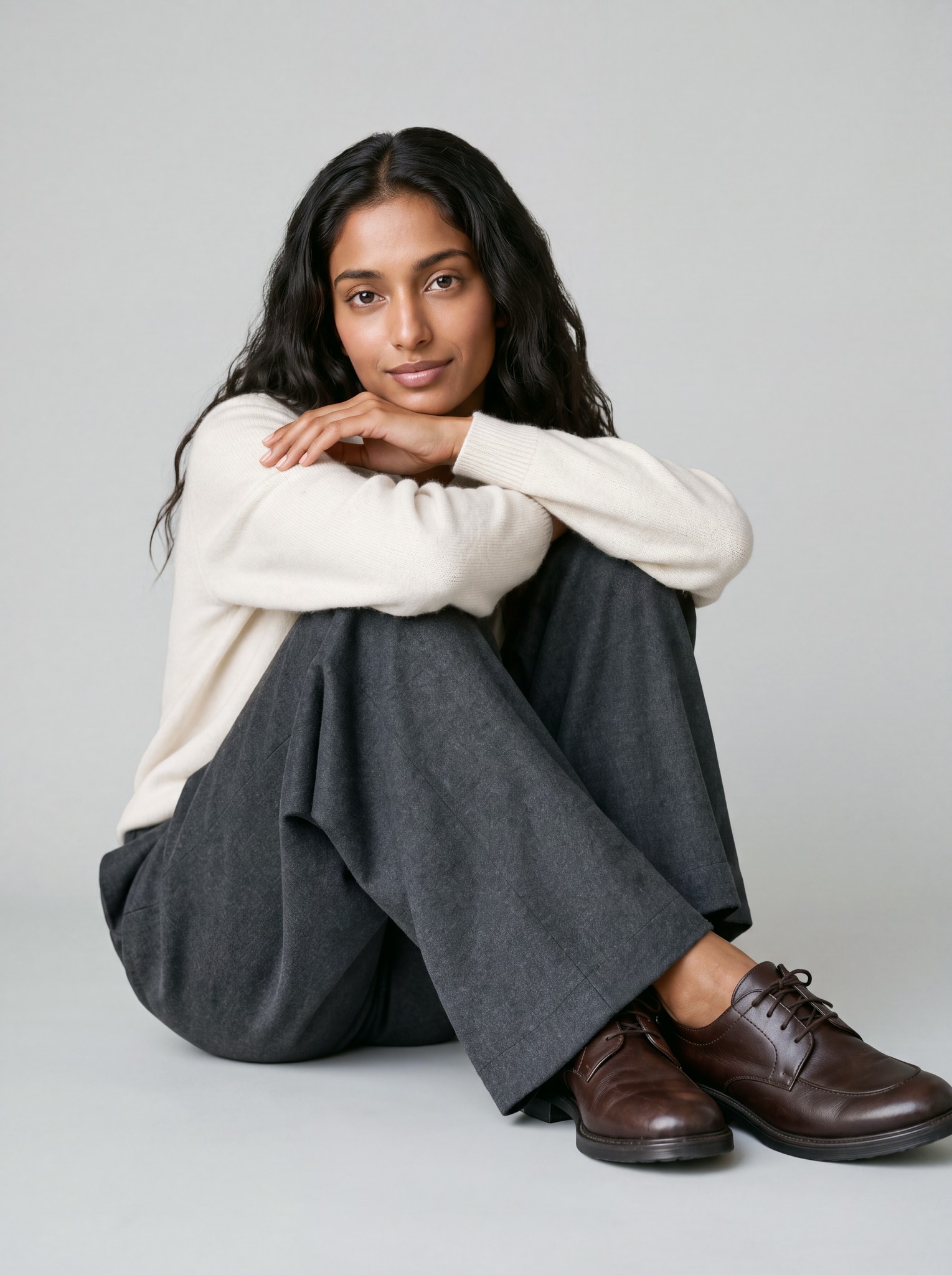A model sitting on a grey studio floor in a cream sweater, grey wide-leg trousers and brown shoes